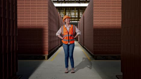 full body of asian female engineer wearing safety helmet looking around while standing with arms akimbo with red brick packed in stacks are stored