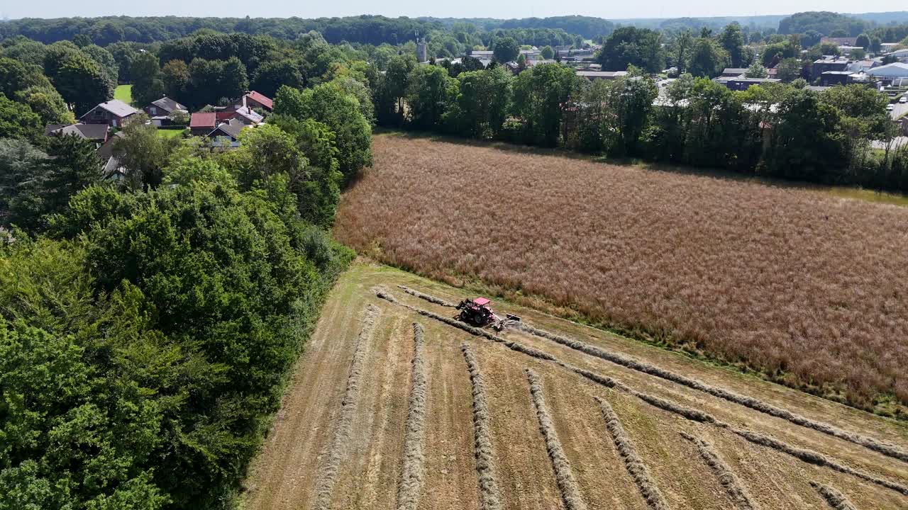 Aerial flyover tractor harvesting golden field near village,in rural district of German town. Agriculture and countryside theme. Top down. Suburb town with working machinery on field in summer