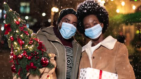 Close-up-view-of-joyful-African-American-couple-wearing-facial-masks-holding-a-Christmas-tree-and-smiling-at-camera-while-it¬¥s-snowing-on-the-street-in-Christmas