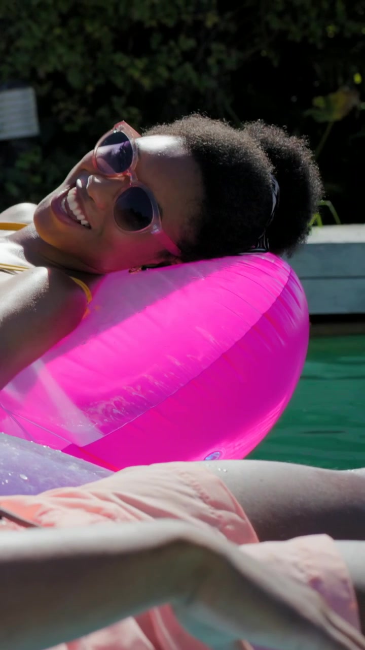 Vertical video: Smiling woman moving head upward resting as cam pans in pool, in suit on pink tube