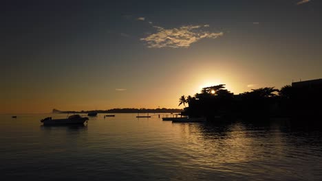 Ein-Langsames-Schwenken-Eines-Kleinen-Piers-Kurz-Vor-Sonnenaufgang,-Während-Die-Sonne-Hinter-Einigen-Palmen-Aufgeht-Und-Kleine-Boote-In-Der-Mitte-Des-Bodens-Vor-Anker-Liegen