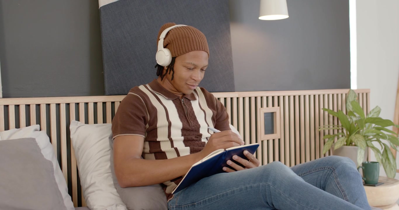Voksen mann sitter på sengen, hvite hodetelefoner sklir, justerer og skriver i en blå notatbok