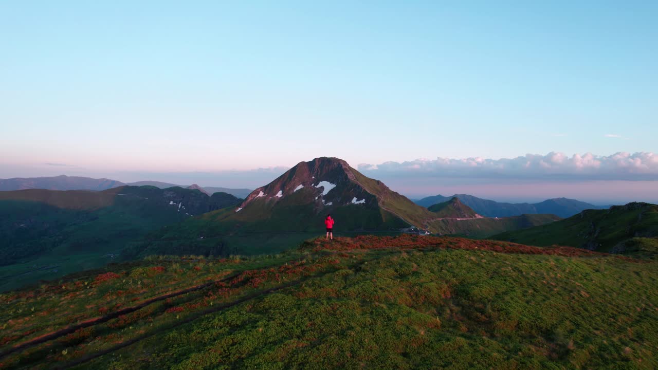 aerial shot revealing Puy Mary Volcano at sunset with a person in red coat looking at the landscape in the foreground from Puy de la Tourte, Cantal departement, Auvergne Rhone Alpes region, France