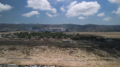 drone moving from the haifa beach to the city as a road cross busy with vehicles, with pleasant blue sky with distant mountains