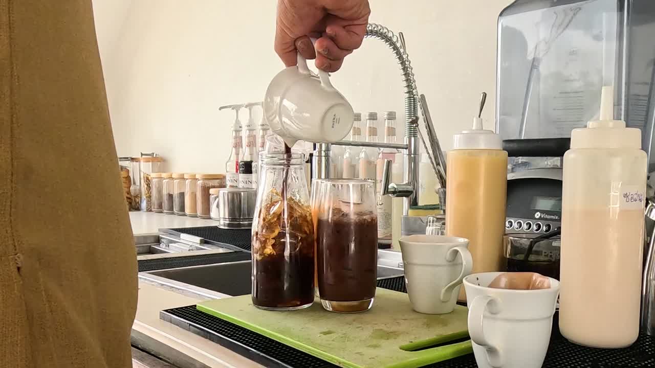 A person pours cream into iced coffee glasses on a kitchen countertop.