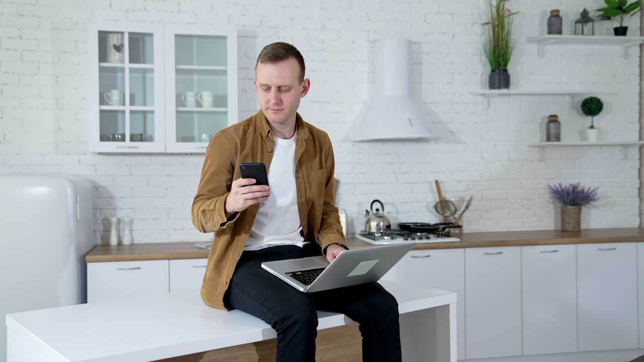 Freelance man working at home. Serious young man sitting on the kitchen table with a laptop and taking photos of himself on a mobile phone.