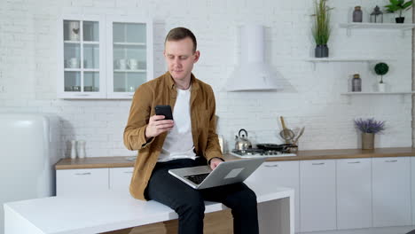 freelance man working at home. serious young man sitting on the kitchen table with a laptop and taking photos of himself on a mobile phone.