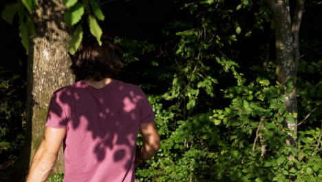 Junger-Mann-In-Rotem-T-shirt-Und-Langen-Haaren-Geht-An-Einem-Sonnigen-Sommertag-In-Slowmotion-In-Einen-Grünen-Dunklen-Wald