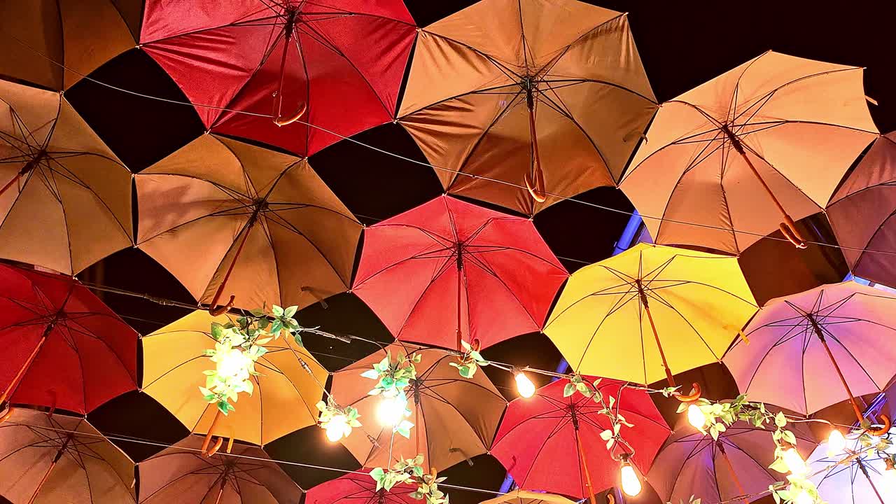 Multiple colourful lit umbrellas hang from wires above street at night