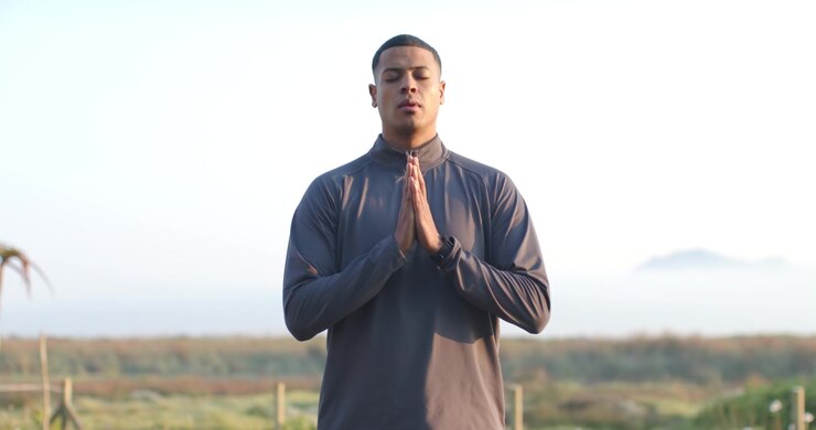 Camera rising, African American man in sportswear meditating with hands at chest on deck by pool