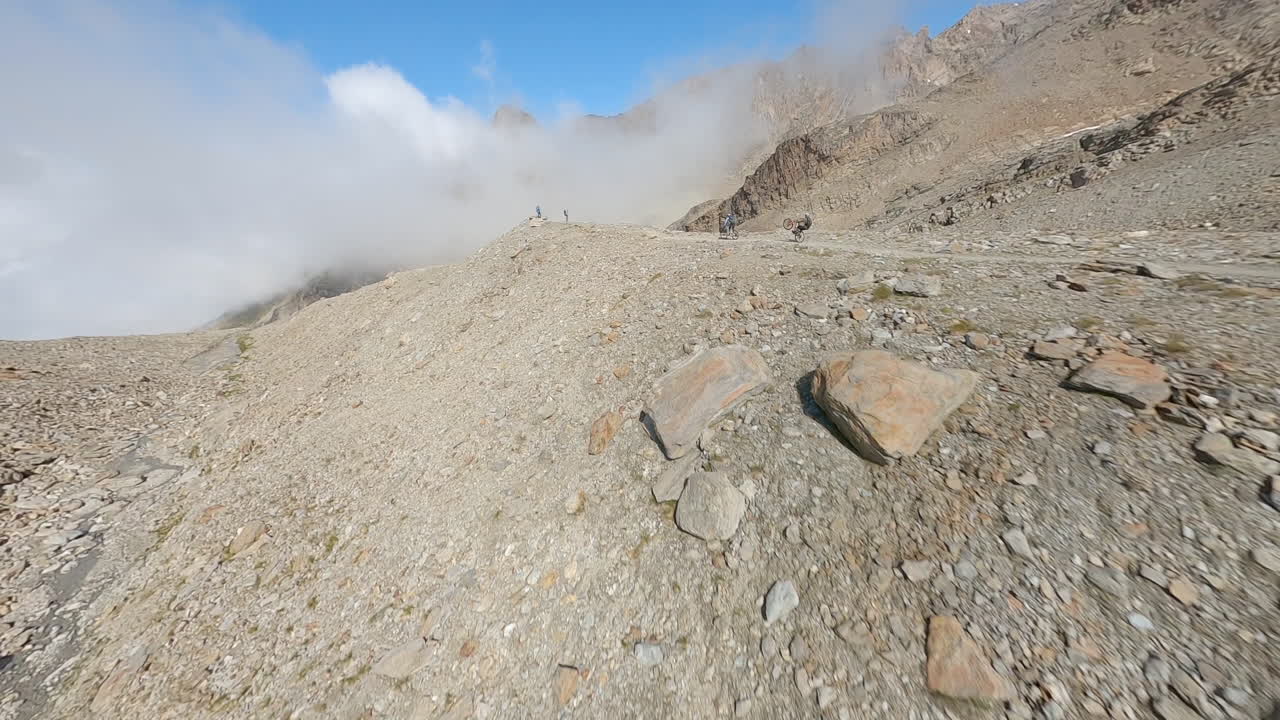 FPV drone follows two mountain bikers descending steep, exposed alpine trails above Saas-Grund and Saas-Fee on a clear bluebird summer day, showcasing epic terrain and speed