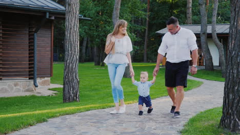 Eine-Junge-Familie-Mit-Einem-Kleinen-Sohn-Geht-Um-Ihr-Haus-Herum,-Sie-Halten-Das-Baby-An-Der-Hand,-Gehen-Al-Walk