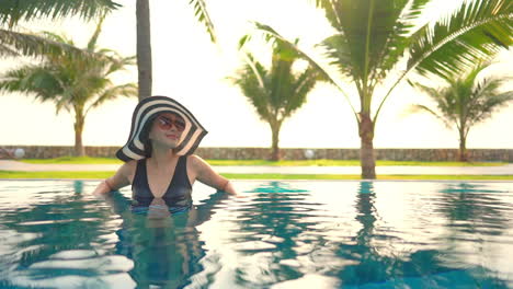 Una-Mujer-Asiática-Sonriente-Y-Feliz-Con-Un-Gran-Sombrero-Blanco-Y-Negro-Dentro-De-La-Piscina-Disfrutando-De-Unas-Vacaciones-Tropicales-Apoyada-En-El-Borde-De-La-Piscina-Al-Atardecer