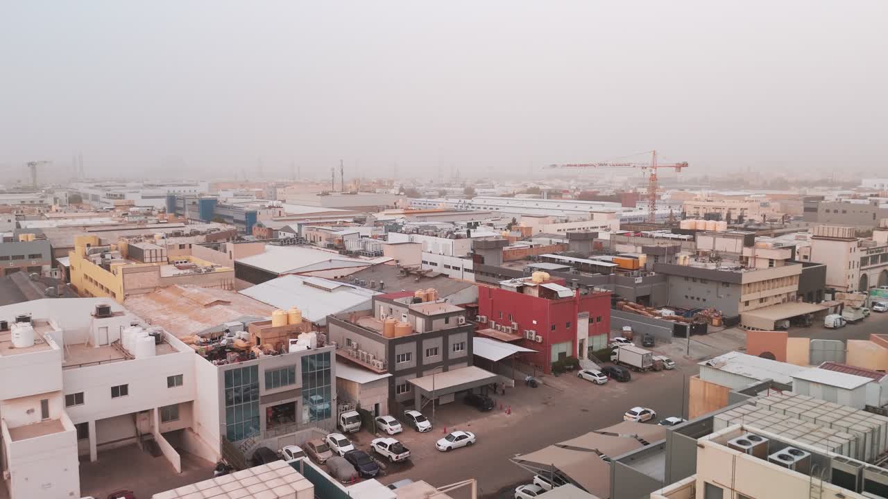 Aerial drone shot of an industrial district with warehouses, rooftops, cranes, and parked vehicles under dusty haze and low visibility in daylight, creating a muted urban atmosphere