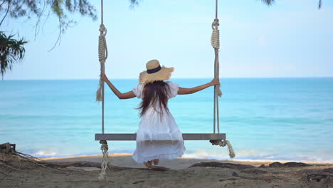 Vista-Trasera-De-Una-Chica-Con-Un-Gran-Sombrero-De-Paja-Y-Un-Vestido-Blanco-En-Un-Columpio-Frente-Al-Mar