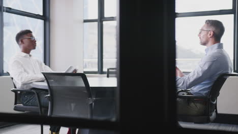 A-black-and-a-white-businessman-talking-in-a-meeting-room,-full-length,-arc-shot,-through-glass-wall