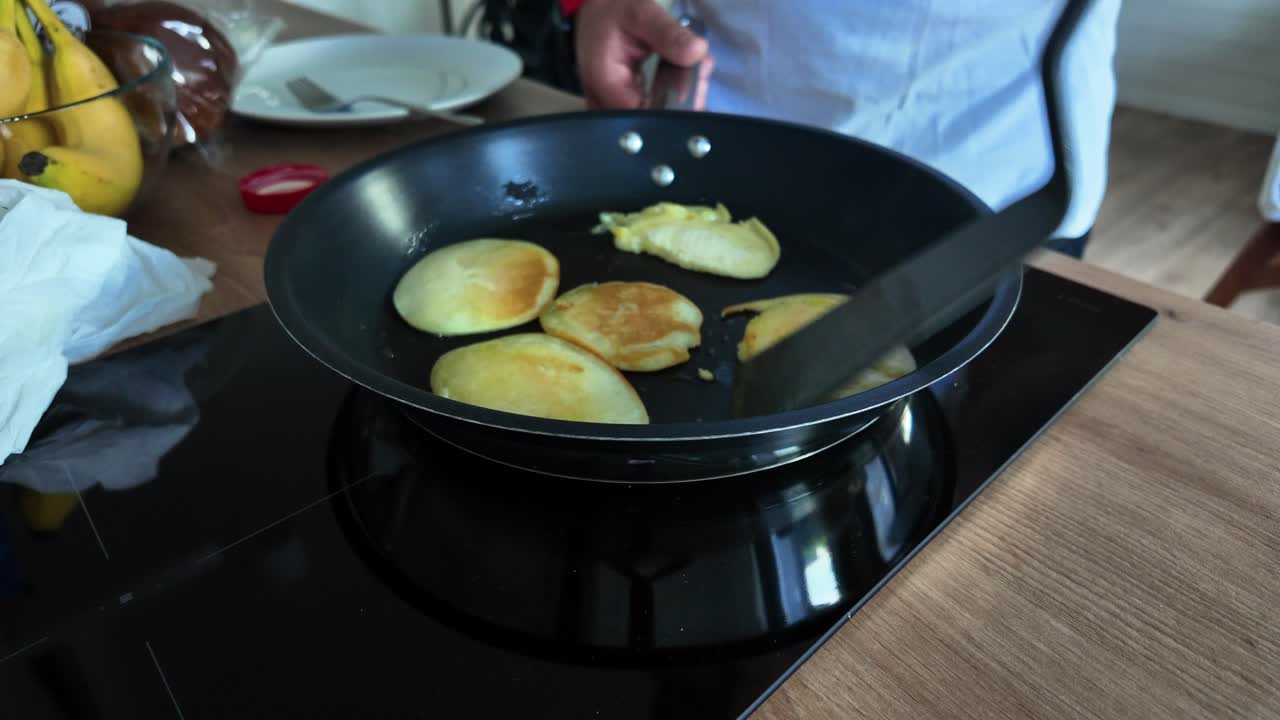 Person flipping golden pancakes in a non-stick frying pan on a modern stovetop in a cozy kitchen setting. Ideal for food, cooking, or breakfast-related content
