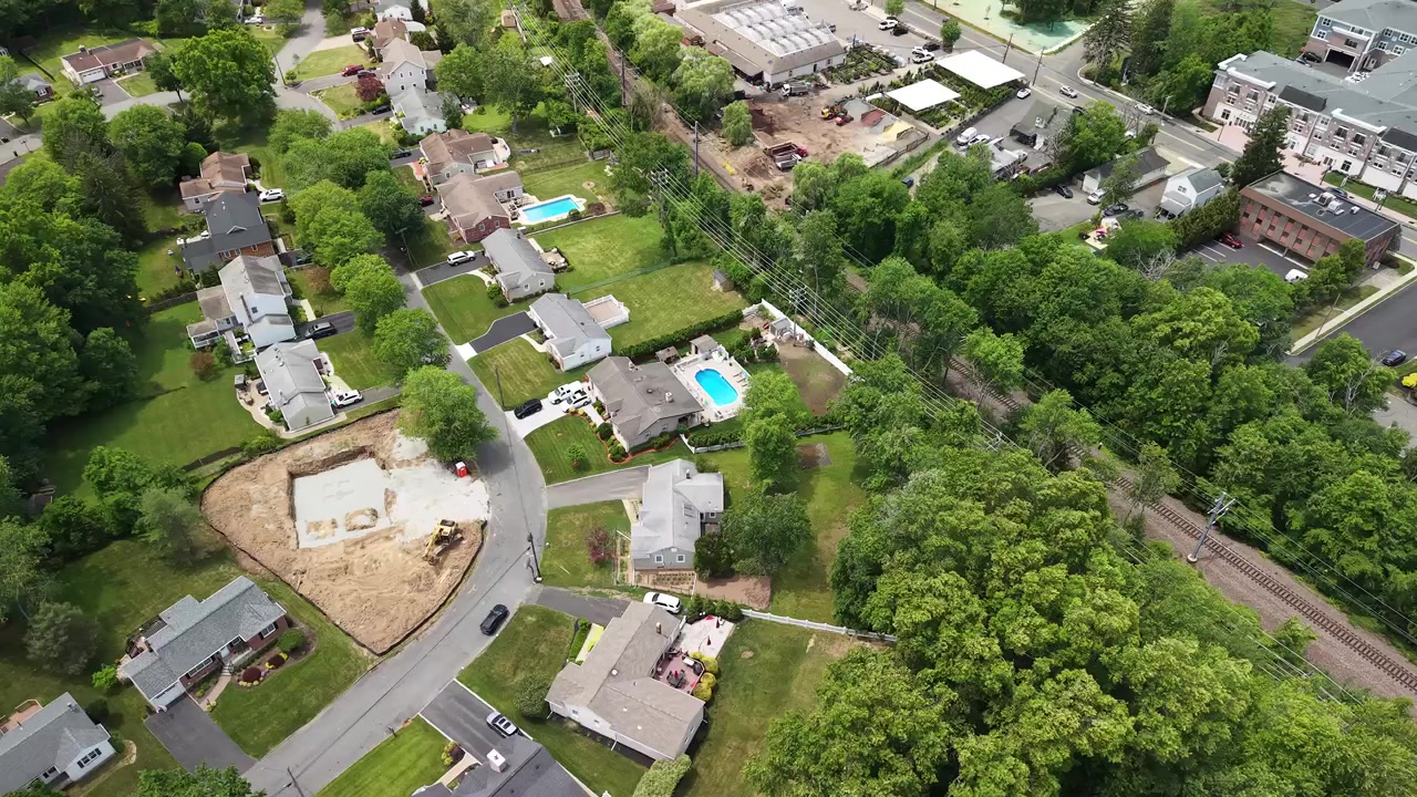 Overhead drone footage flying above a suburban american town, showing residential houses with pools, green trees, a construction site, and adjacent railroad tracks during a sunny summer day