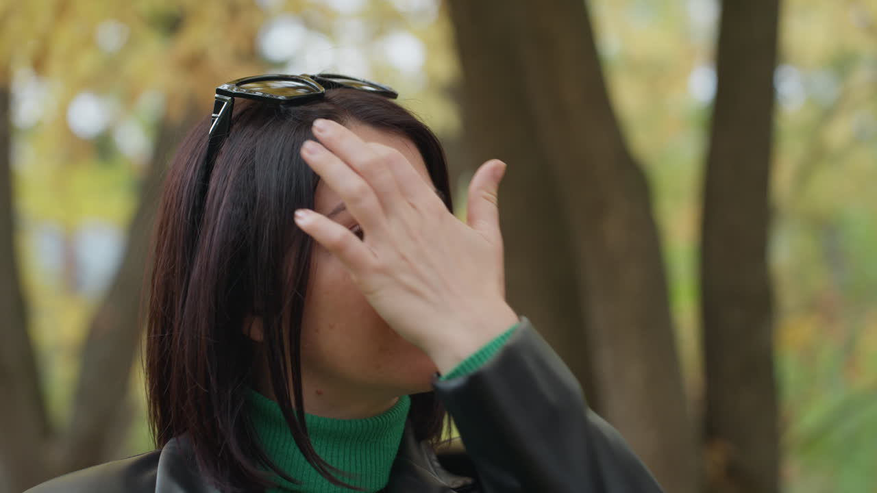 woman portrait leather jacket autumn park, contemplative woman adjusts hair with sunglasses on head, wearing green turtleneck and black leather jacket, standing among golden trees with soft natural