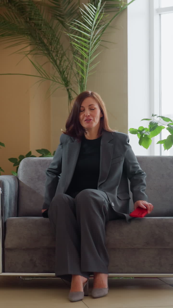 Confident businesswoman in elegant gray suit standing beside sofa in stylish modern office lobby holding red smartphone surrounded by indoor plants natural sunlight professional setting