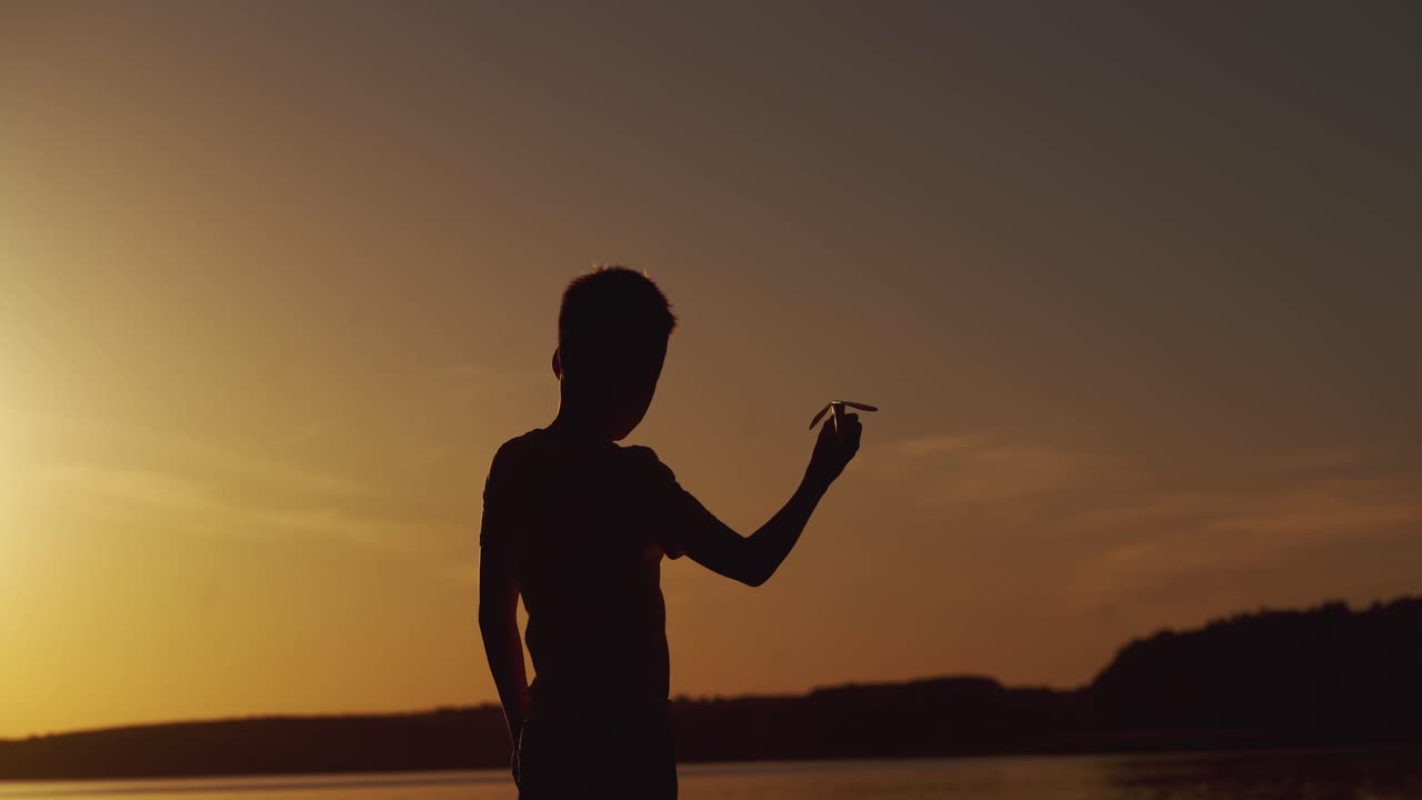 Silhouette of little boy sitting alone at the river on sunset background. Happy child playing with paper airplane.