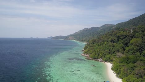 Aerial-view-of-lush-island-with-clear-waters-and-coral-reefs-by-beach-in-Thailand---camera-pedestal-up-tilting-down