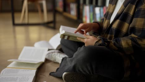 primer plano de una estudiante con una camisa a cuadros leyendo un libro mientras está sentada en el suelo entre los libros de la biblioteca