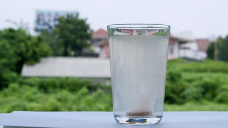 agua burbujeante en un vaso causada por la dilución de un antiácido o bicarbonato de sodio en un telón de fondo de casas y vegetación verde fuera de foco