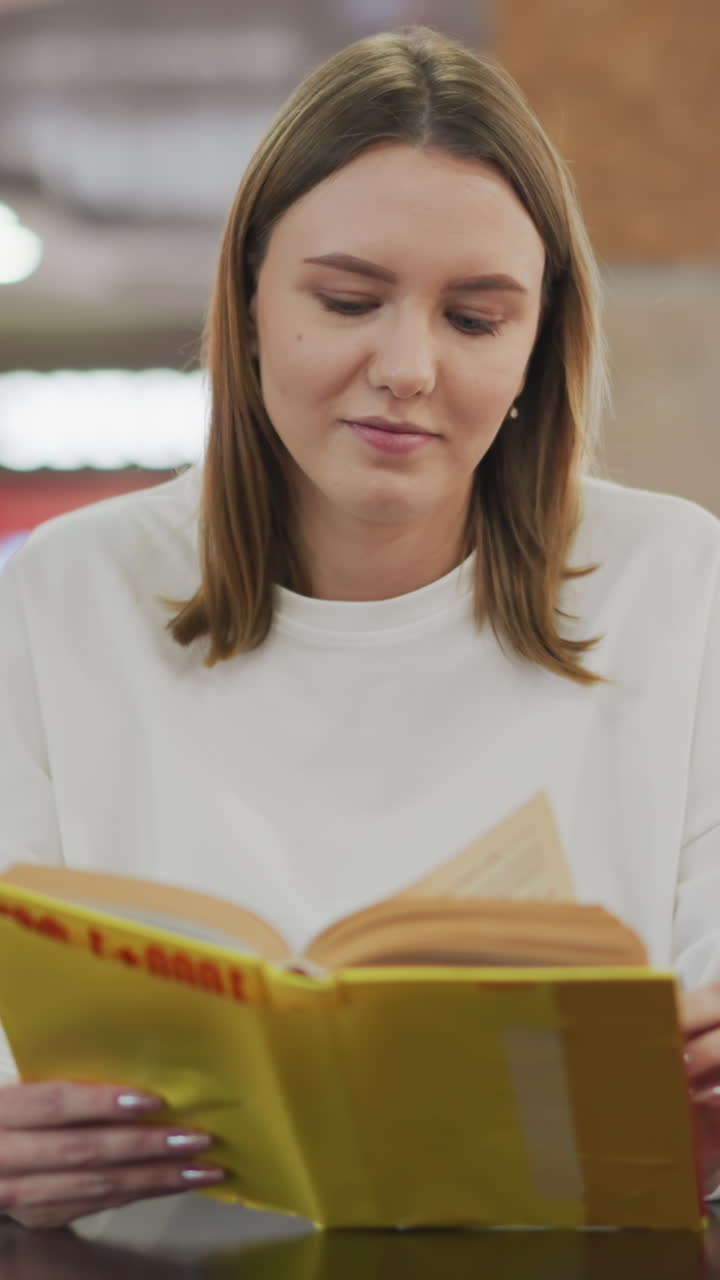 joven leyendo un libro amarillo brillante con una sonrisa sutil en un restaurante moderno, rodeado de un fondo borroso vibrante, con una vista borrosa de otros compradores