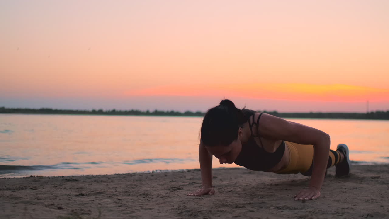chica deportiva haciendo flexiones en la playa al amanecer. joven en forma bronceada chica sonriente con camiseta deportiva y leggings haciendo flexiones en la playa al atardecer