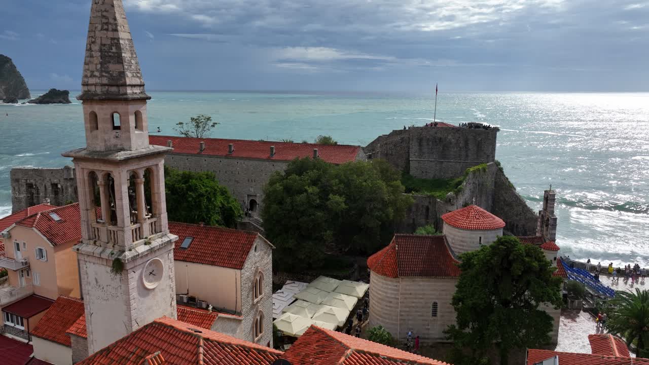 Tracking left across old tower spires in Budva with dramatic sky and waves near waterfront town walls