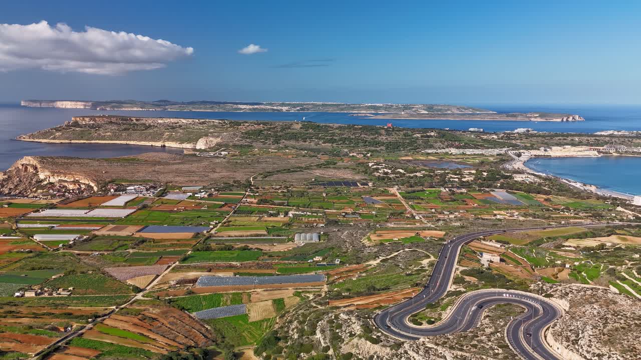 Stupefacente hyperlapse aereo di Mellieha, Malta, che cattura la sua costa panoramica, i rigogliosi campi agricoli e il Mar Mediterraneo sotto un cielo azzurro limpido con nuvole sparse.