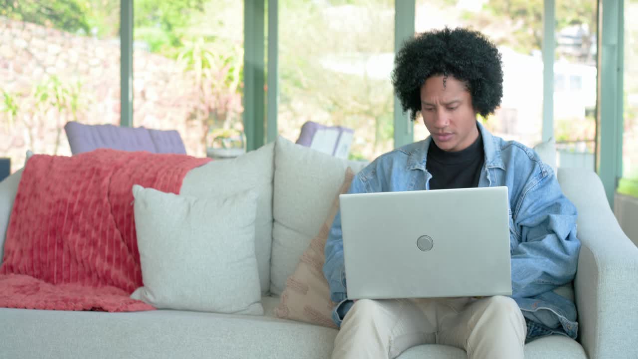 Mid adult man opening silver laptop beside window, typing while walking to sofa and continuing work