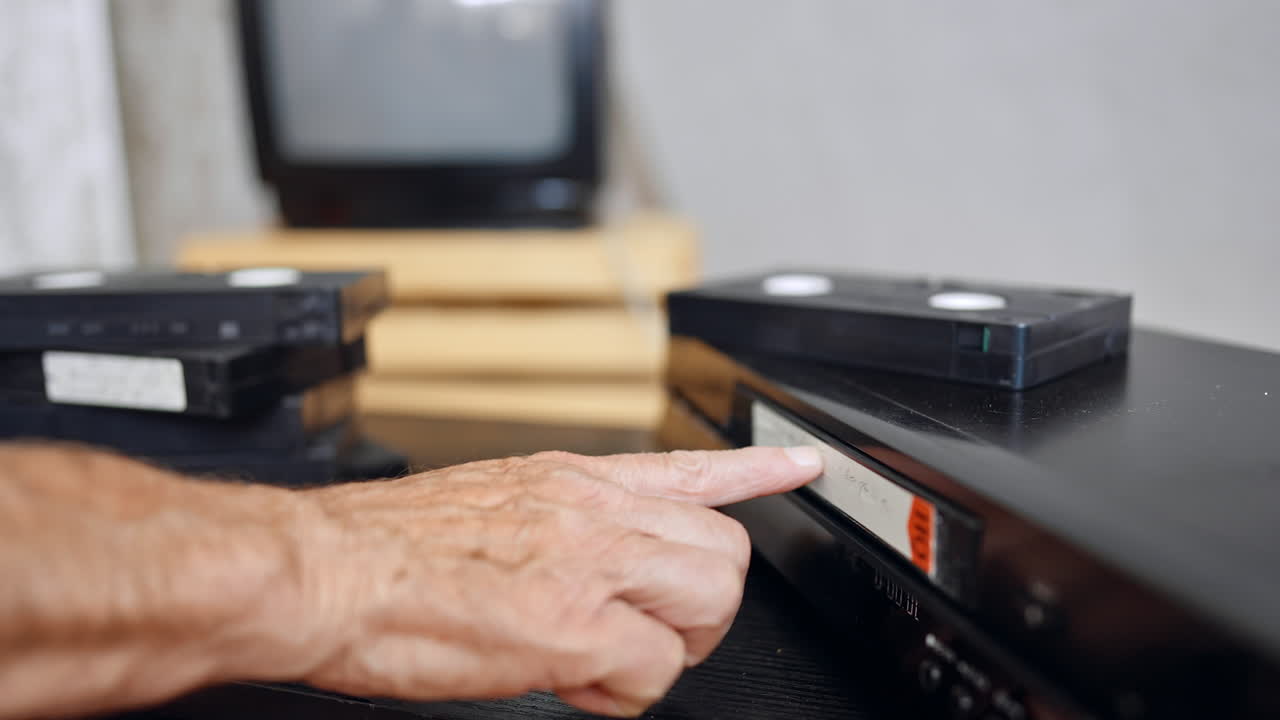 Hand guiding VHS into player. A person pushes a VHS tape into an old video recorder with other cassettes nearby