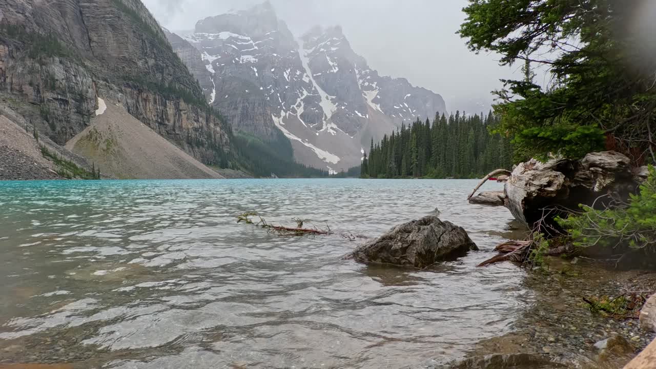 Moraine lake with slightly choppy waters and mountains in the background on a foggy and rainy summer day