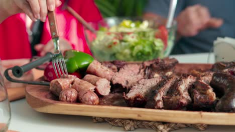 closeup of several people's hands holding forks while skewering pieces of roast meat served on a wooden board on a table with a bowl of salad outdoors during daytime