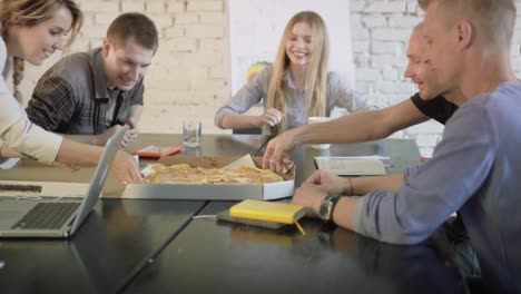 young creative coworkers have a meal together. business team are eating pizza in the office. woman brings a box of pizza to office workers and they are happy