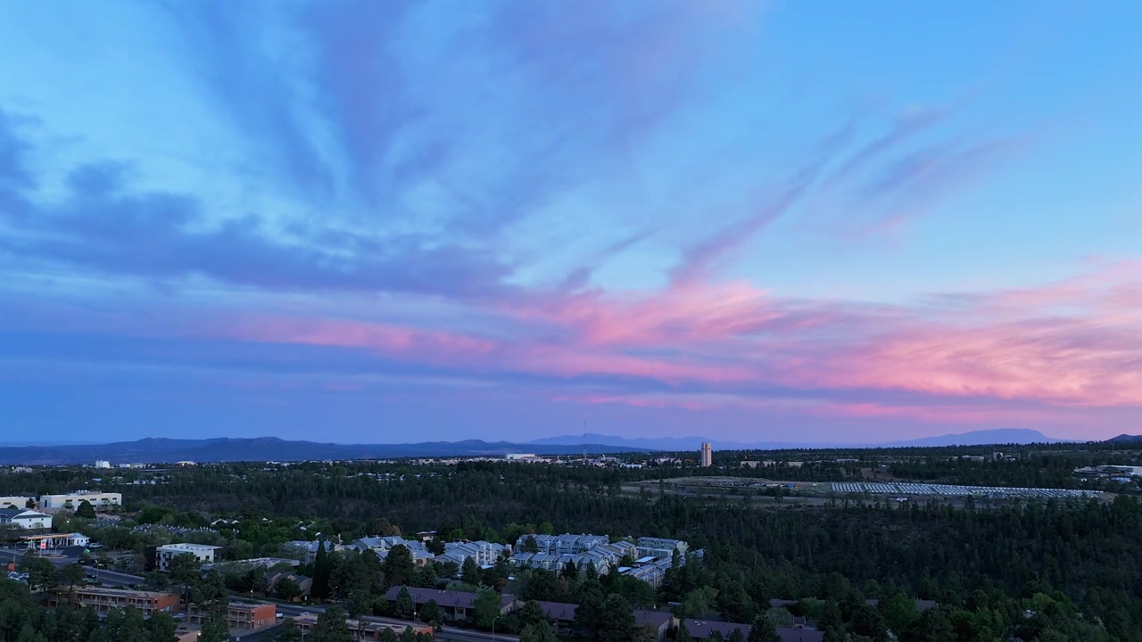 Sidelengs time-lapse droneopptak fra luften over Los Alamos, New Mexico ved solnedgang. Himmelen forvandles idet dynamiske skyer skifter fra gylne nyanser til dype kveldslilla toner over den vidstrakte forstadsbyen