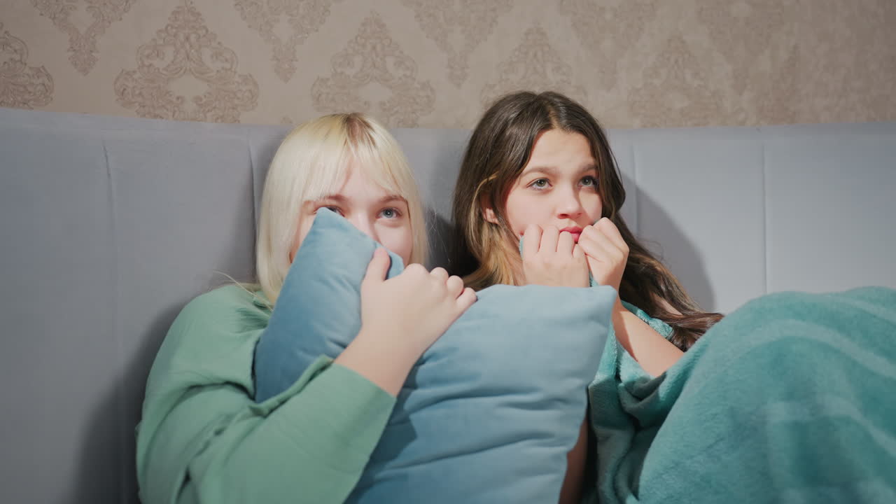 two young women sitting close in bed wrapped in blanket, watching scary movie with frightened expressions, one clutching pillow and other biting blanket