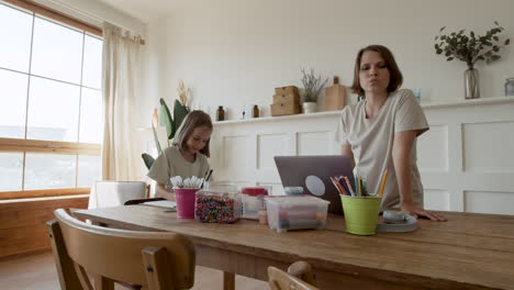 a mother gives her blonde daughter pencils so she can draw while she gets ready to work from home on her laptop