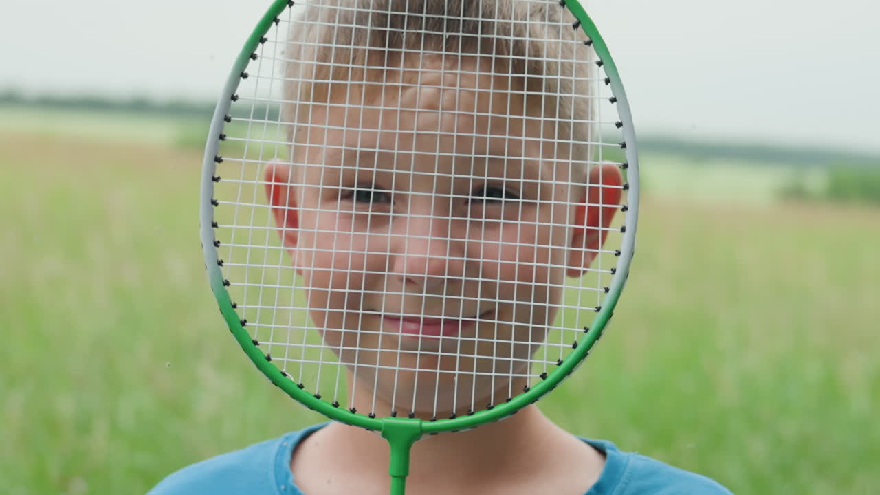 배드민턴 라켓을 들고 초원에서 웃고 있는 백인 소년, 햇빛이 비치는 풀과 멀리 보이는 나무가 있는 솔직한 야외 인물 사진, 편안한 여름 분위기와 활기찬 에너지, 시골에서 포착한 활기찬 어린 시절