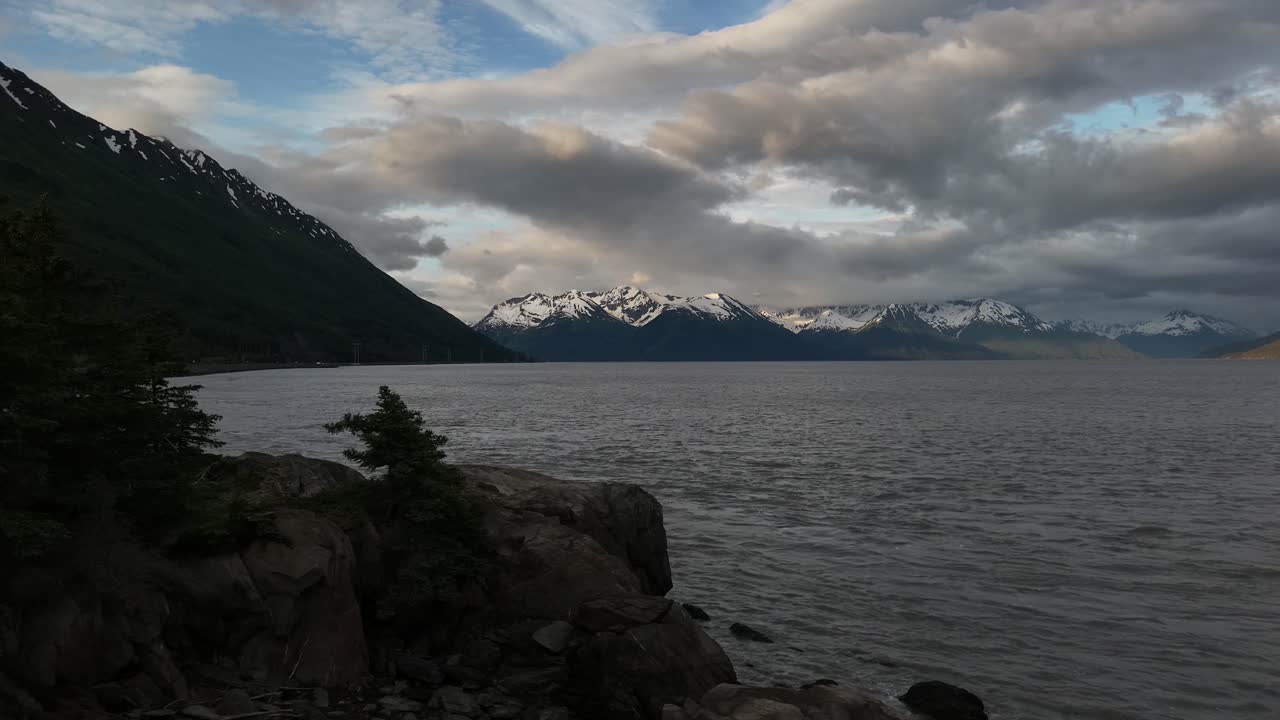 Inlet of water with Snowcapped mountains in background, partly cloudy sky with patches of blue, trees and boulders in foreground, Turnagain Arm, Kenai Mountains, Late June in Alaska