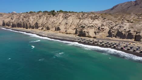 Aerial-panning-across-Greek-Island,-rocky-cliff-mountains-and-blue-water-waves-in-the-Mediterranean-Sea-on-white-beach-in-Santorini,-Greece