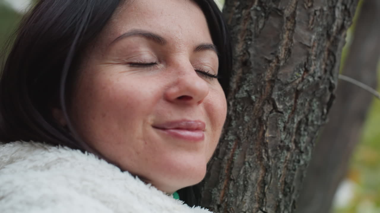 Donna bianca appoggiata a un albero che sorride a occhi chiusi, respira l'aria della foresta, tiene in mano una tazza calda, pennello con foglie autunnali, una morbida coperta sulle spalle, espressione calma, momento di radicamento e cura di sé