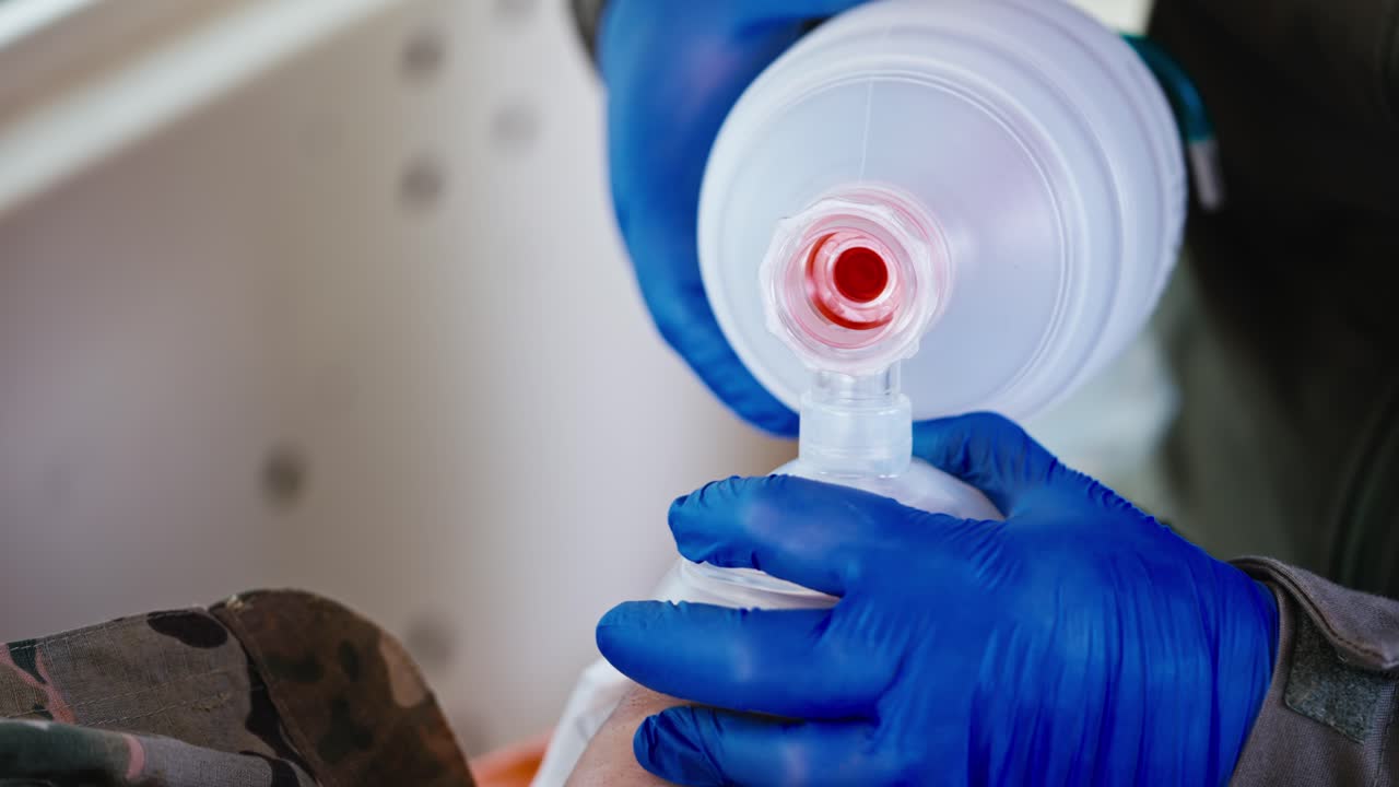 Close up a confident male military medic in blue rubber gloves pumps air using a matte white bottle and performs artificial respiration on an unconscious wounded soldier in an ambulance