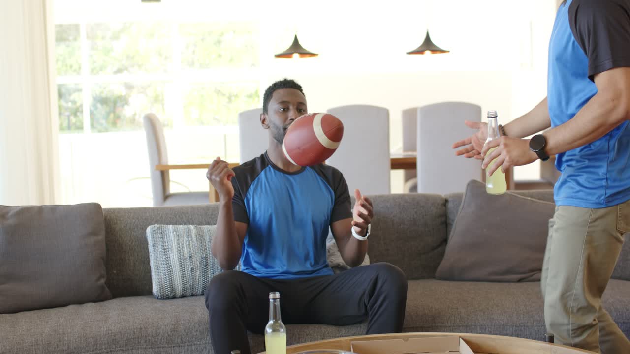 Diverse male friends catching flying football on couch, friend offering drink reacting to screen