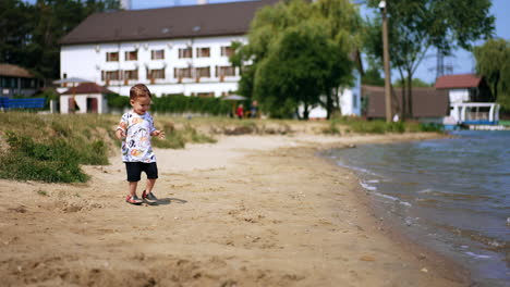 lovely caucasian baby standing on the shore. little boy takes some sand to play with. summer time joy.