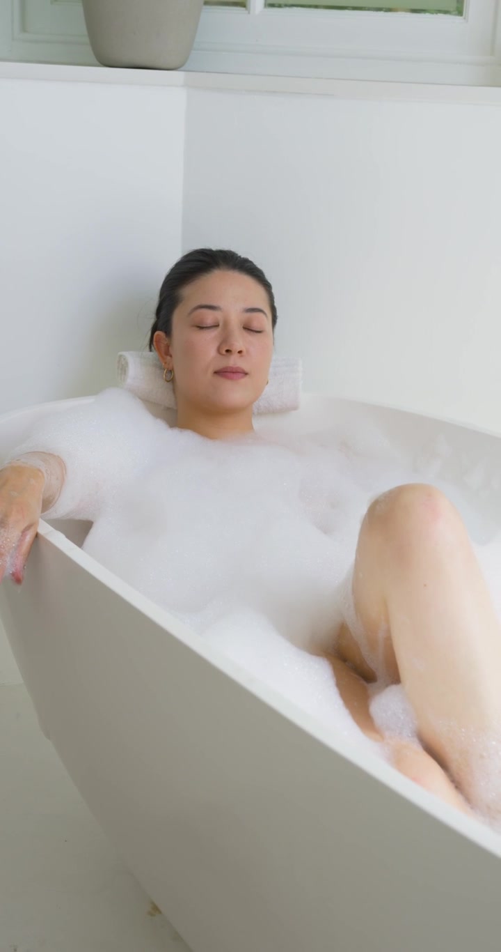 Woman reclining in bathtub, making small leg shift, settling bubble foam for spa wellness