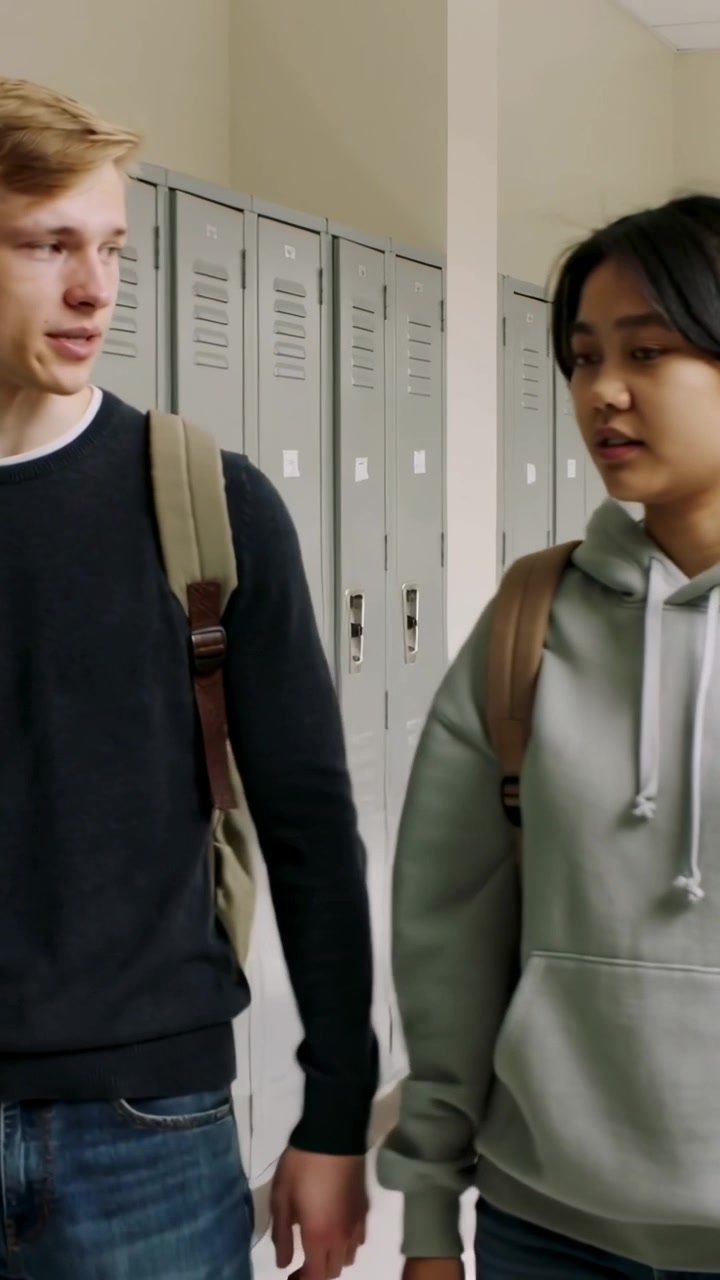 Vertical video: Walking two students talking in school hall, passing lockers and wearing backpacks