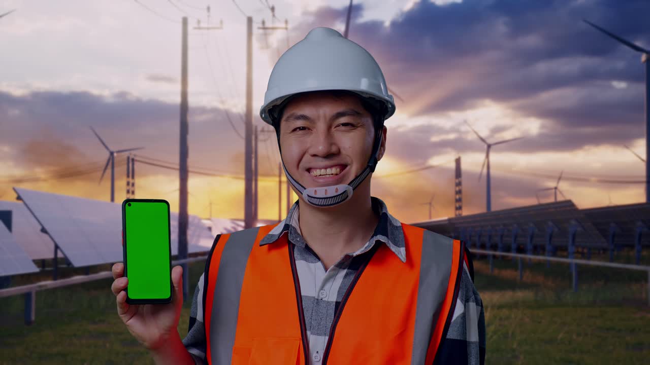 Close Up Of Asian Male Engineer With Safety Helmet Smiling And Showing Green Screen Smartphone To The Camera While Standing With Solar Panel and Wind Turbines